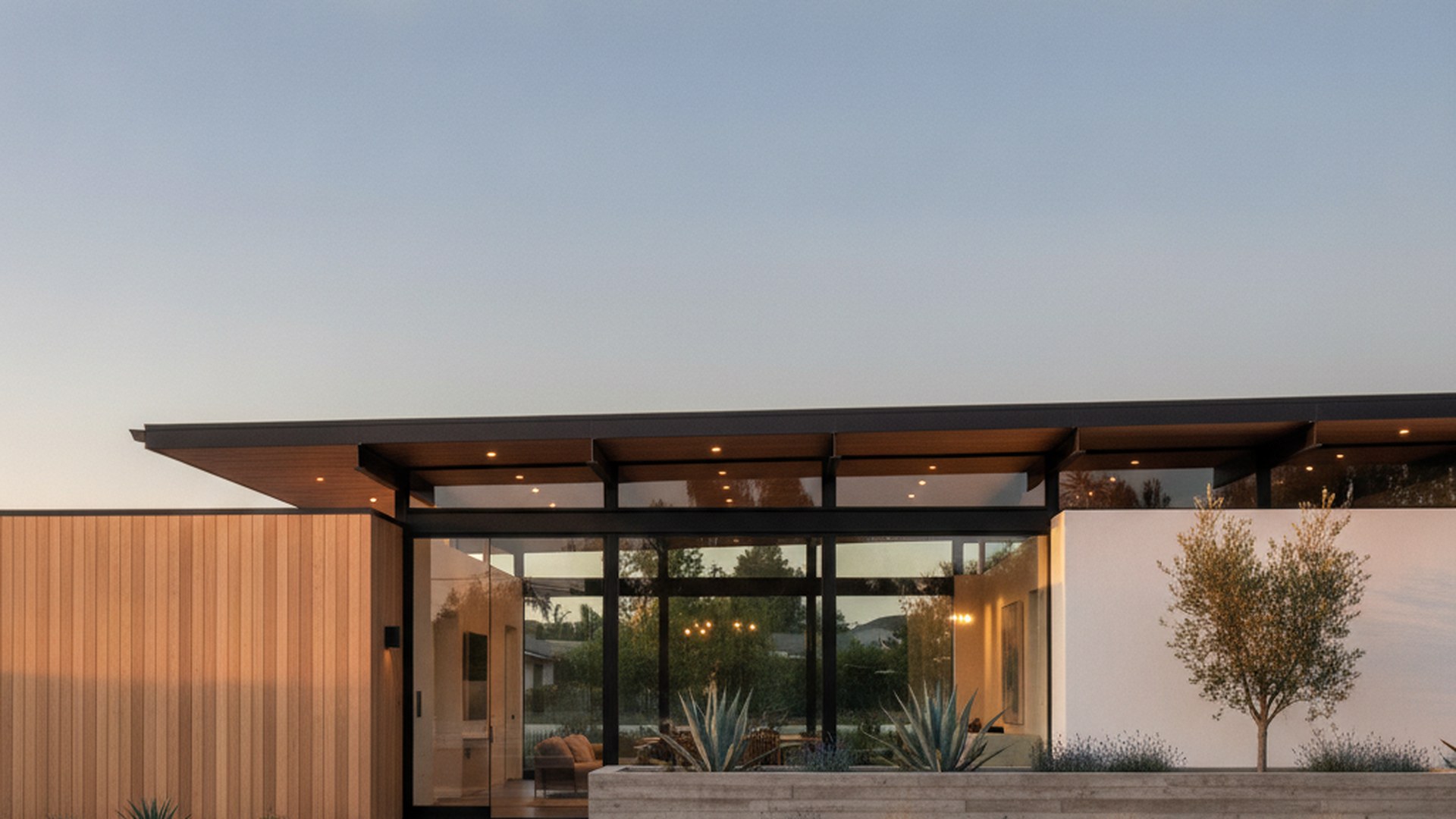 Single-story architecturally modern Orange County home at dusk with a low-slope roof, deep overhangs and walls of glass glowing from within